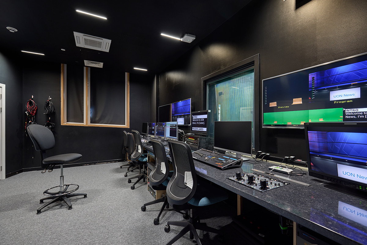 Internal view of the TV Studio Control Room, where walls are painted black and there are desktop screens on a long workbench with different keyboards with various TV studio buttons. There are screens which are showing the studio room view as well as a glass window, which leads to the studio.