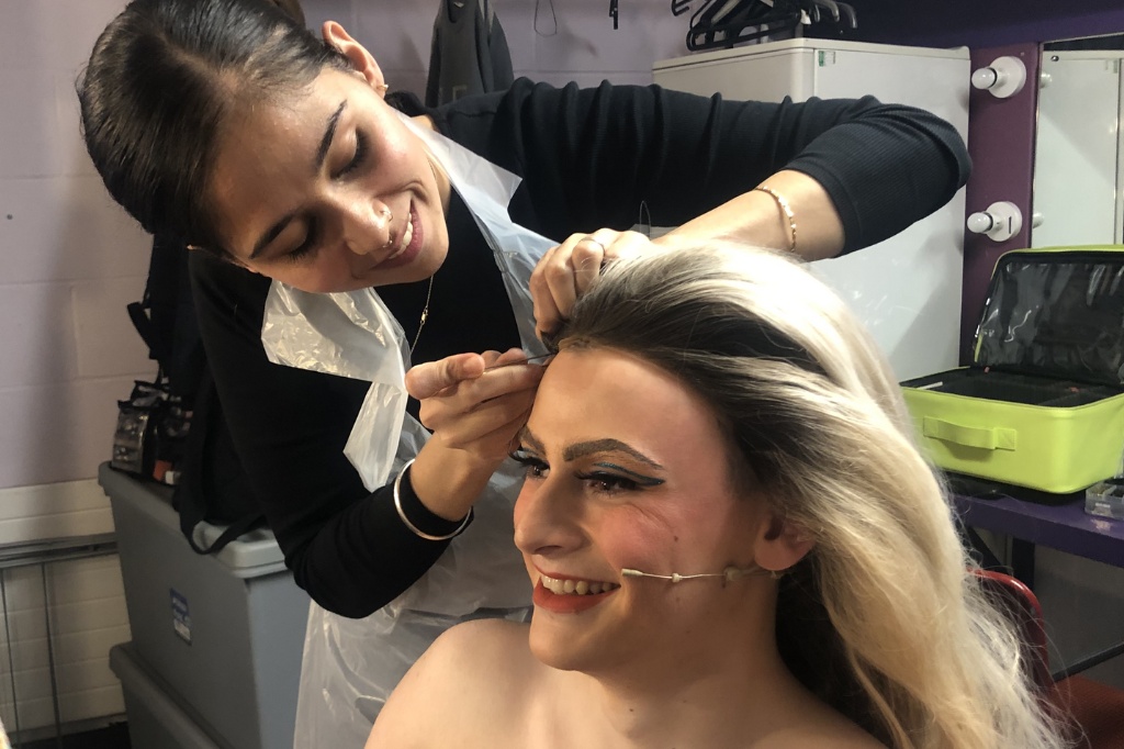 Student places makeup on actor who smiles in mirror.