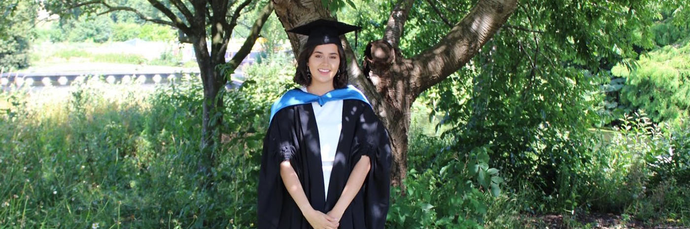 A photo of Nina Garner, who is a University of Northampton Geography graduate, wearing her black and blue graduation robes and mortarboard on her graduation. There are trees and plants in the background