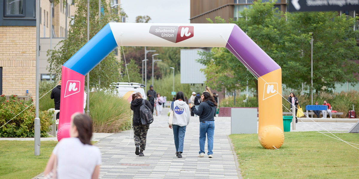 Students walking under Students Union' archway at Welcome Weekend on campus. The archway is colourful with the SU logo and the campus is looking green with trees and has other students around.