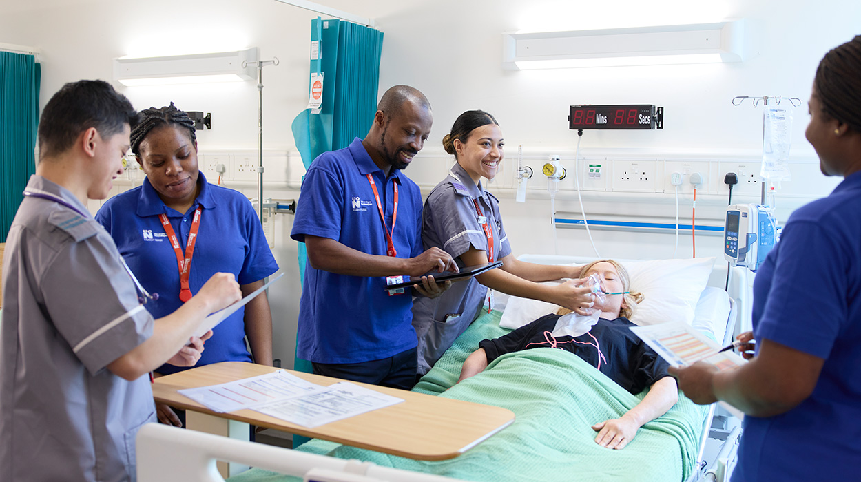 Five Adult Nursing students in a session in a ward, one student holding an oxygen mask on a student lying in the bed.