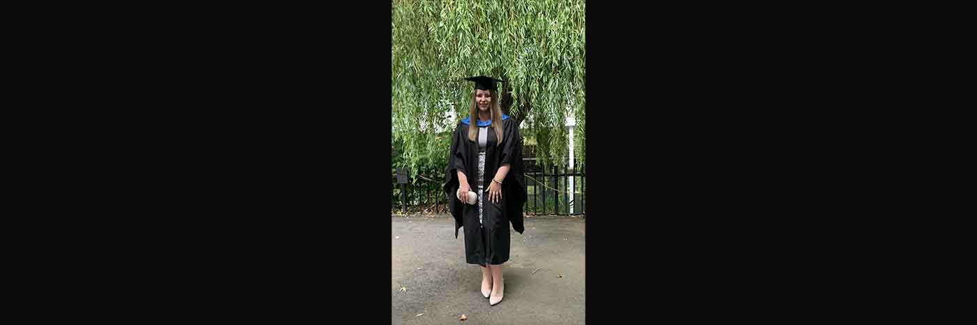 The photo shows recent UON graduate Aimee Fearns who graduated in 2021 with a Geography BSc (Hons) degree. Aimee is wearing her official graduation robe and mortarboard at her graduation ceremony, and posing for the graduation photo in front of a large tree and railings