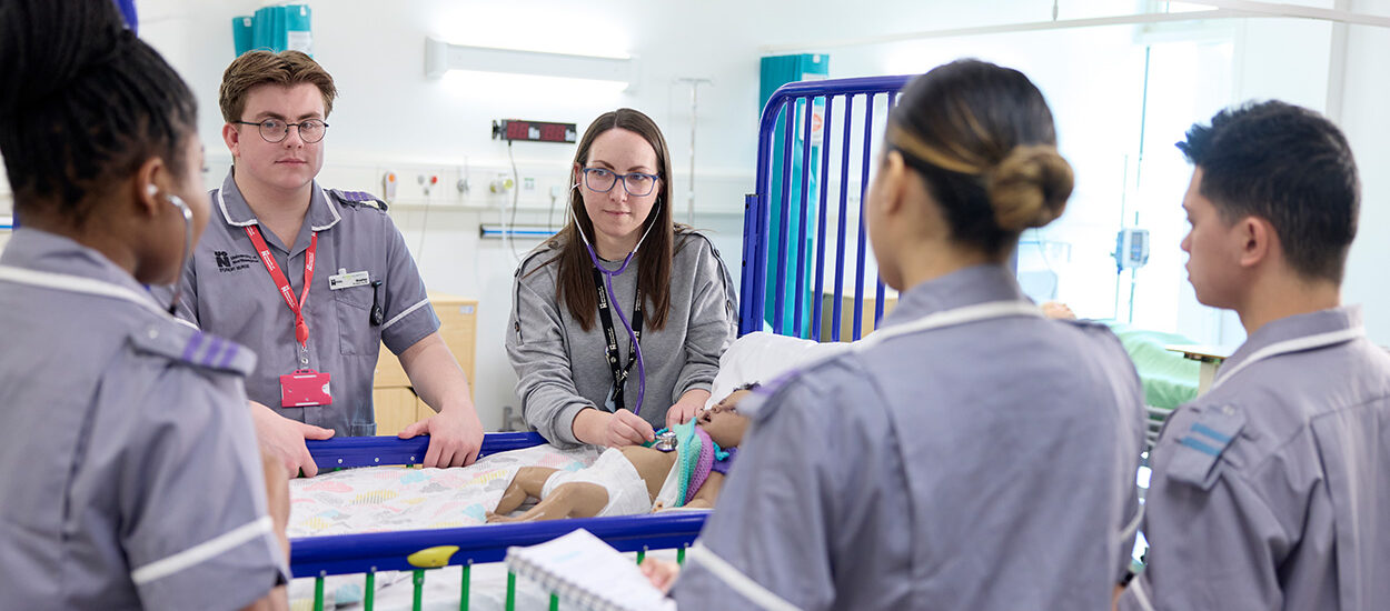 Four students in a demo with an academic holding a stethoscopes on a child manikin.