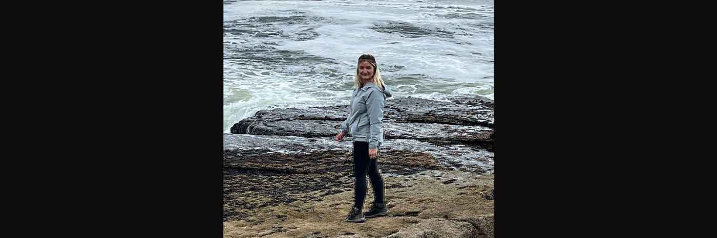 The photo shows Geography BSc graduate Georgia Keenan by the seashore with rocks and waves in the background