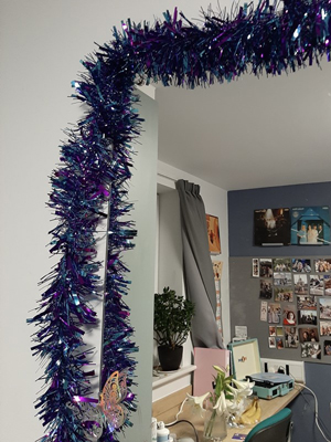 Tinsel around a mirror in halls, in the reflection can be seen a desk with a record player and a vase with white lilies.