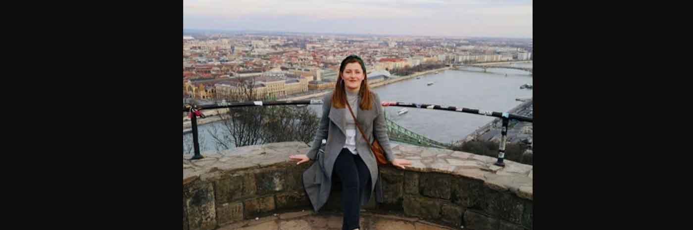 The photo shows Geography BSc graduate Kerrie Plunkett sitting on a low wall with a cityscape behind her