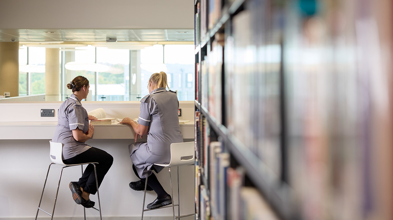 Two students wearing the UON nurse uniform sitting at a high table reading a book in the library.