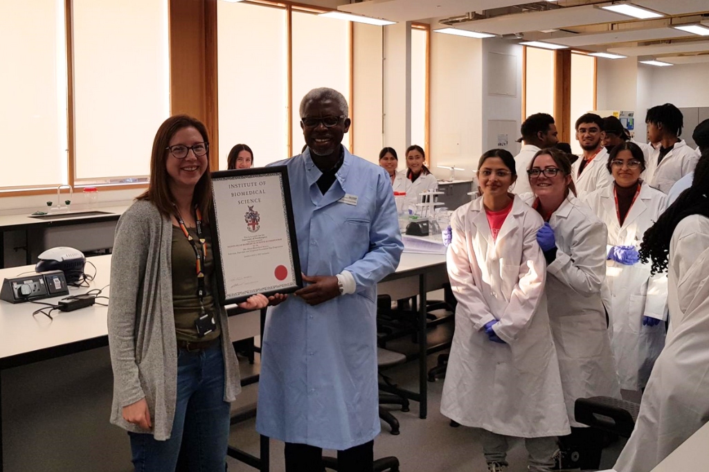 Scientists hold up certificate surrounded by students in white coats.