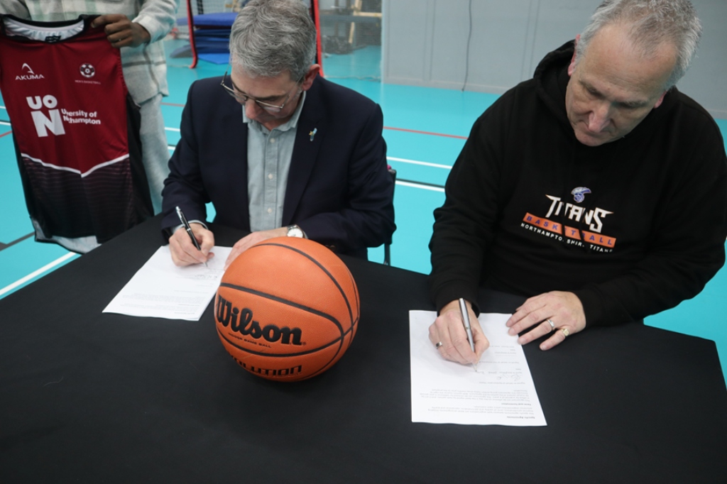 John Sinclair and Pete Burgoine sign sheets of paper on desk next to basketball.