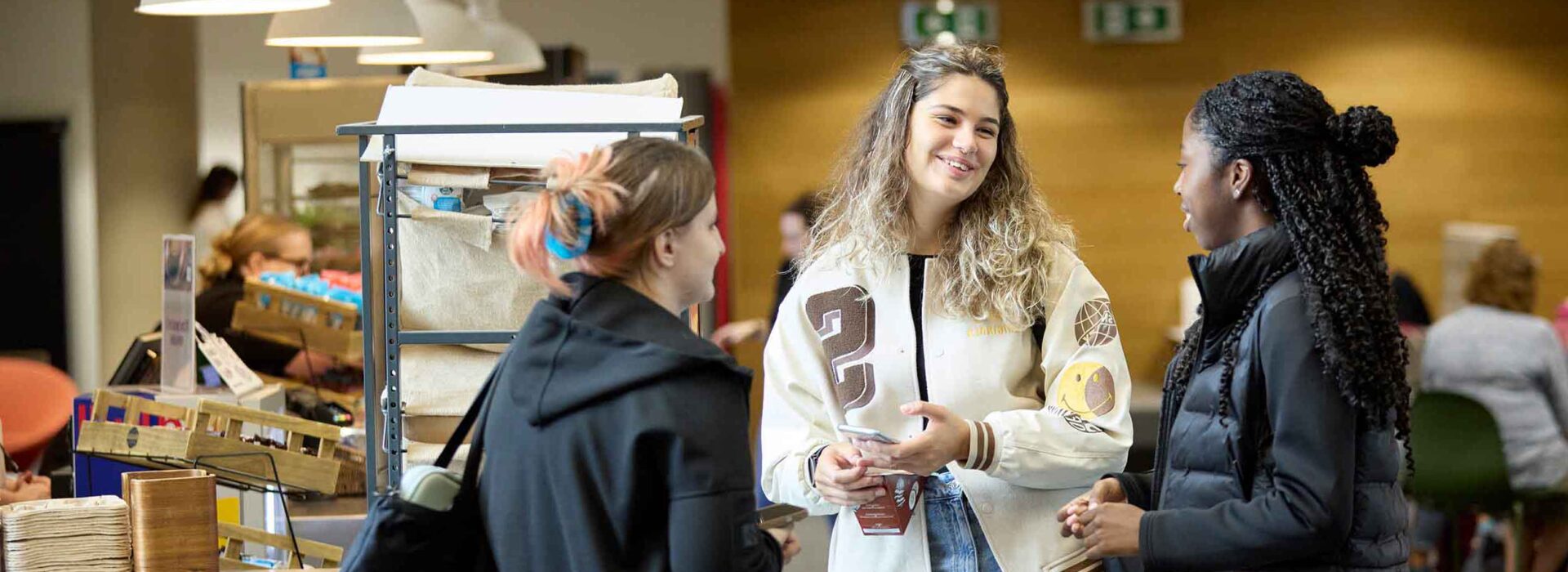 Three students are chatting in the Ground cafe area of the Learning Hub at the University of Northampton. All three students are smiling.