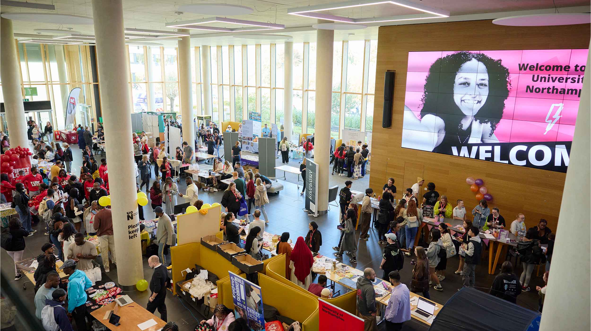 New students visit a Student Union Societies Fair at the University of Northampton. There are approximately 20 advice and information desks - one for each university society. A large display screen on the wall displays a welcome message