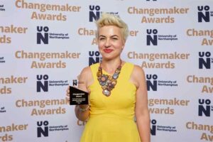 Dr. Tereza Aidonopoulou-Read poses for a photo in front of a photo backdrop at the 2023 UON Changemaker Awards. Tereza holds her award for Education Changemaker of the Year