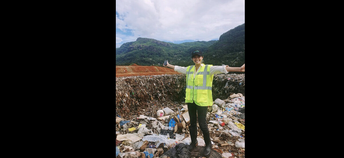 Estelle Patricia Lau Tee in a high-visibility vest standing in front of a landfill with arms outstretched, mountains in the background.