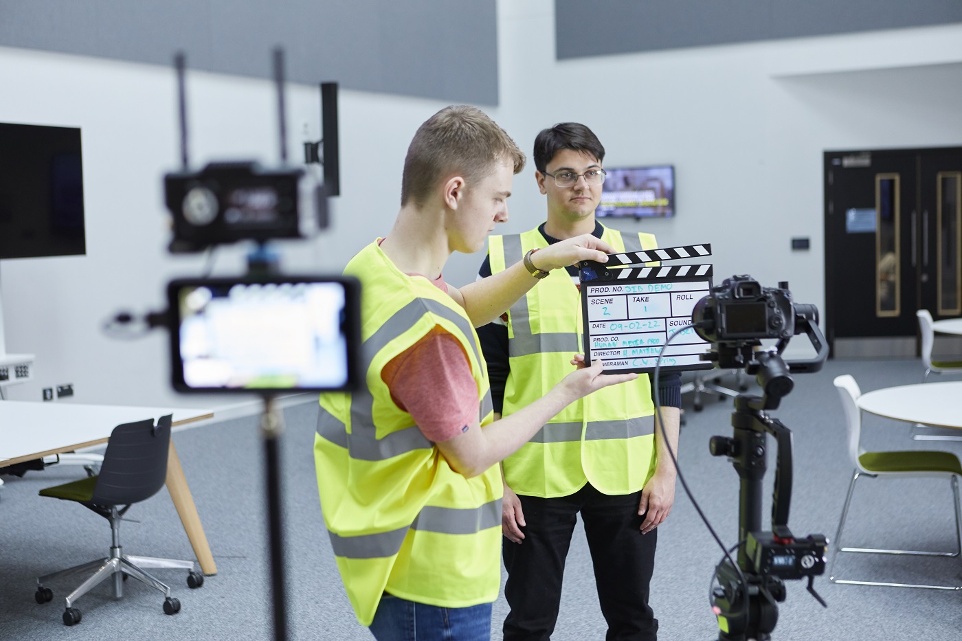 One student holds a clapperboard in front of a camera, while another stands behind.