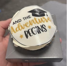A cupcake with white frosting and an edible topper reading "and the adventure begins" in black and gold script, held above a silver box.