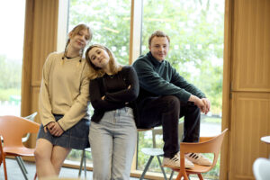 Three students, two standing and one sitting on a chair, pose casually in a well-lit Creative Hub with large windows and orange chairs.