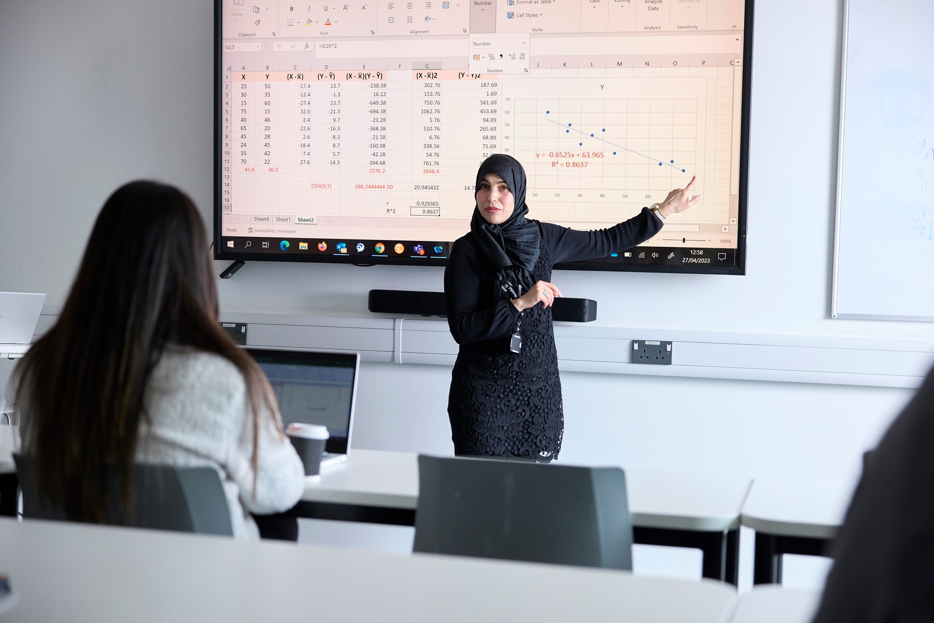 An academic is standing in front of a screen explaining a spreadsheet and table to a student during a class. The student is seated at a table facing the academic with an open laptop