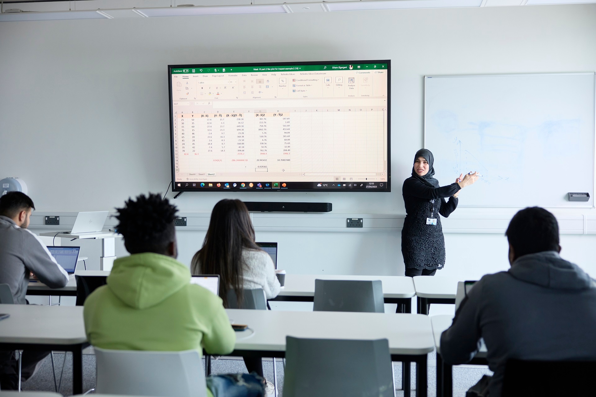 Photo shows an academic standing in front of a screen and a whiteboard and explaining a task to a group of students during a class. The students are seated at tables facing the academic.