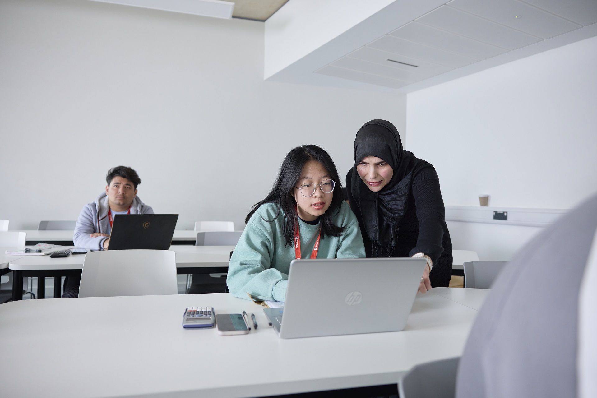 An academic in a black headscarf and a student in a light green sweater are looking at a laptop screen together during a class. Behind them, a student is using their own laptop