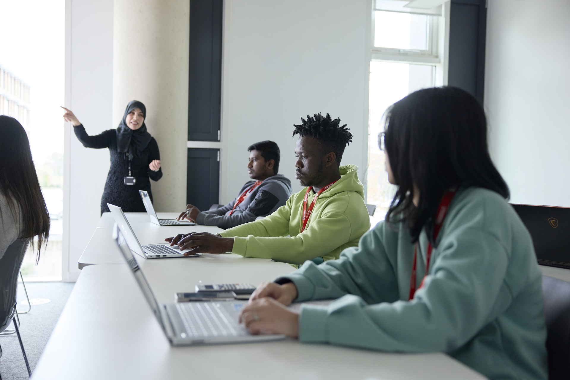 An academic teaches a class while standing in front of three students, who are in the foreground. The students are all using their laptops.