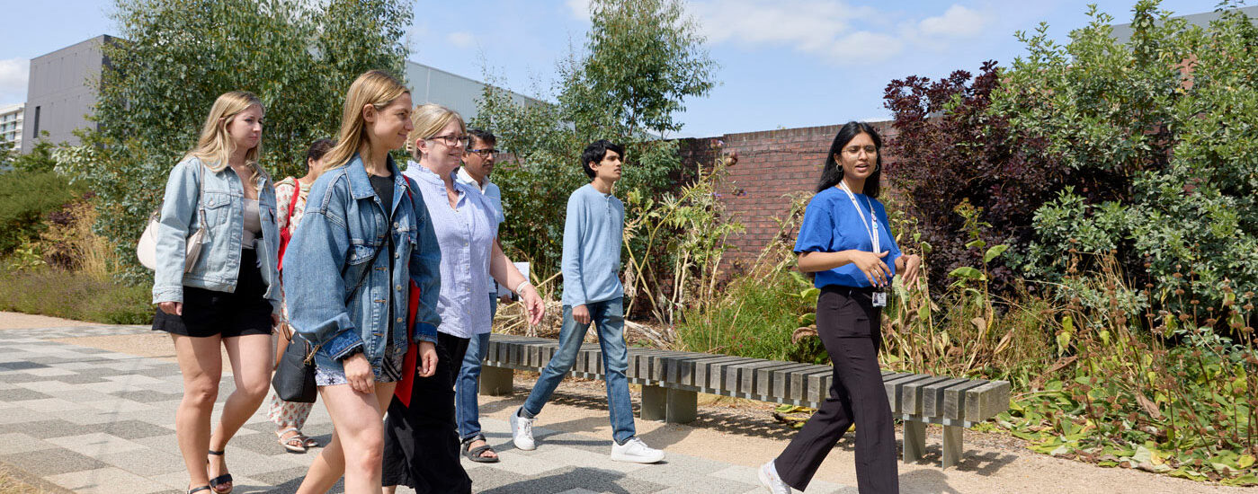 A group of people walking together outdoors on a path with greenery in the background. A student ambassador is leading the group.
