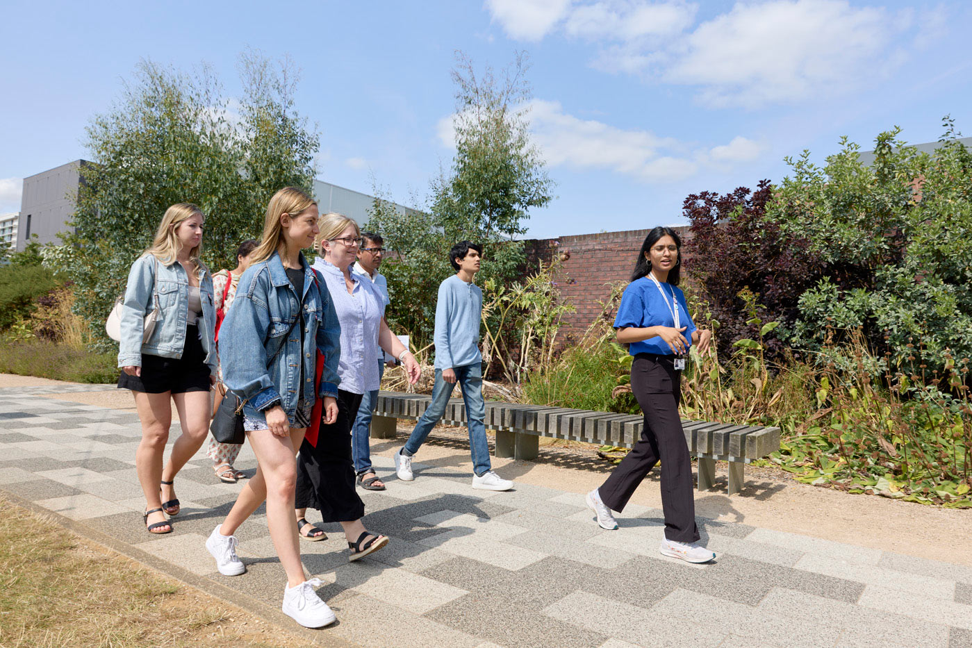 A group of people walking together outdoors on a path with greenery in the background. A student ambassador is leading the group.