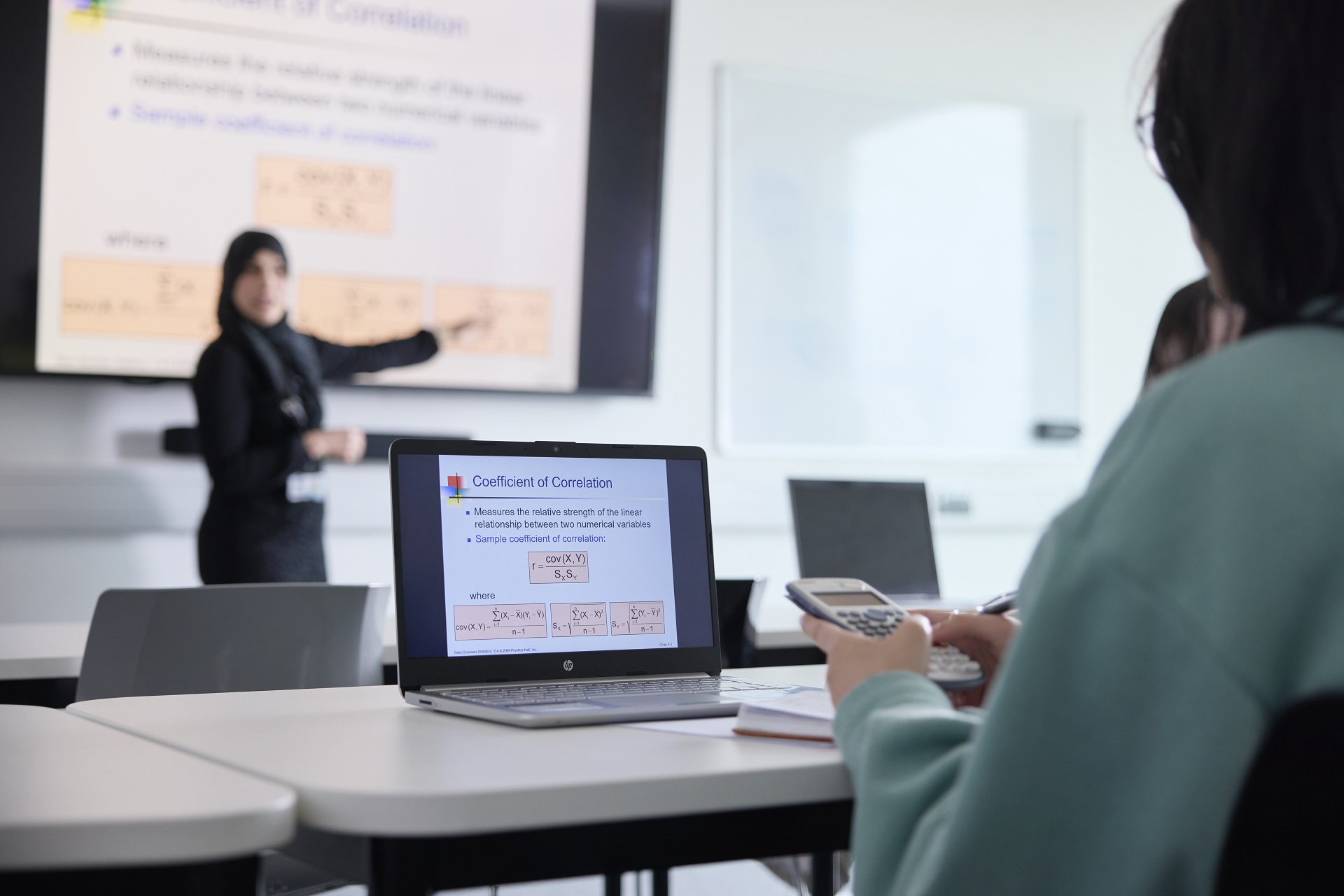 Student using a calculator and looking at their laptop screen, which is displaying module content during a class. In the background, a blurred academic stands in front of a display screen.