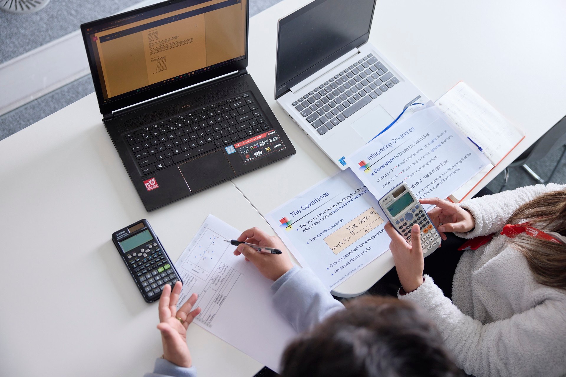 Above view of two students working together on an activity sheet and using calculators and laptops. One student is writing on an activity sheet