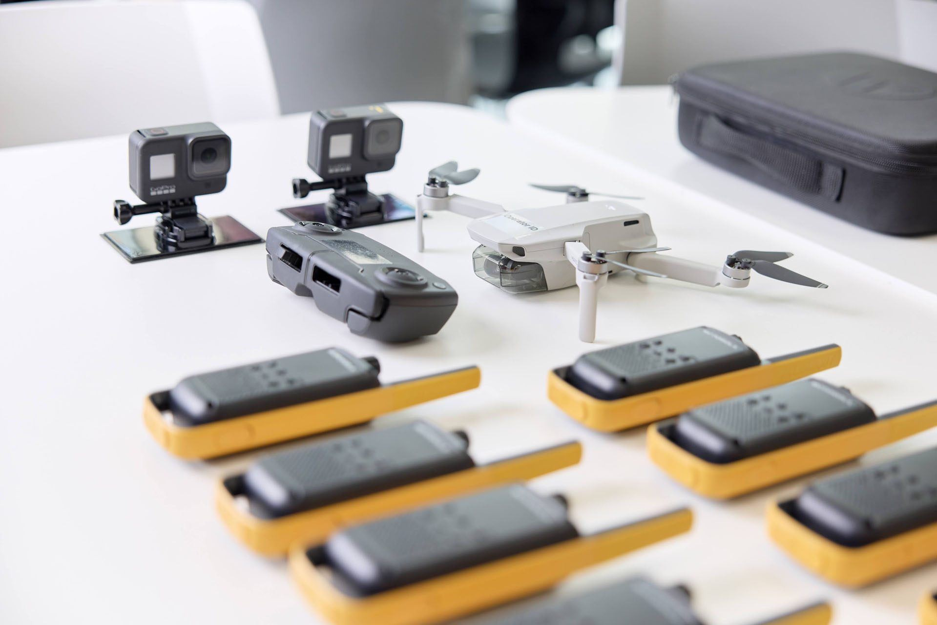 Close-up of surveillance equipment on a table, including cameras, a drone, a controller, and several yellow walkie-talkie radios. In the background is a black carrying case.