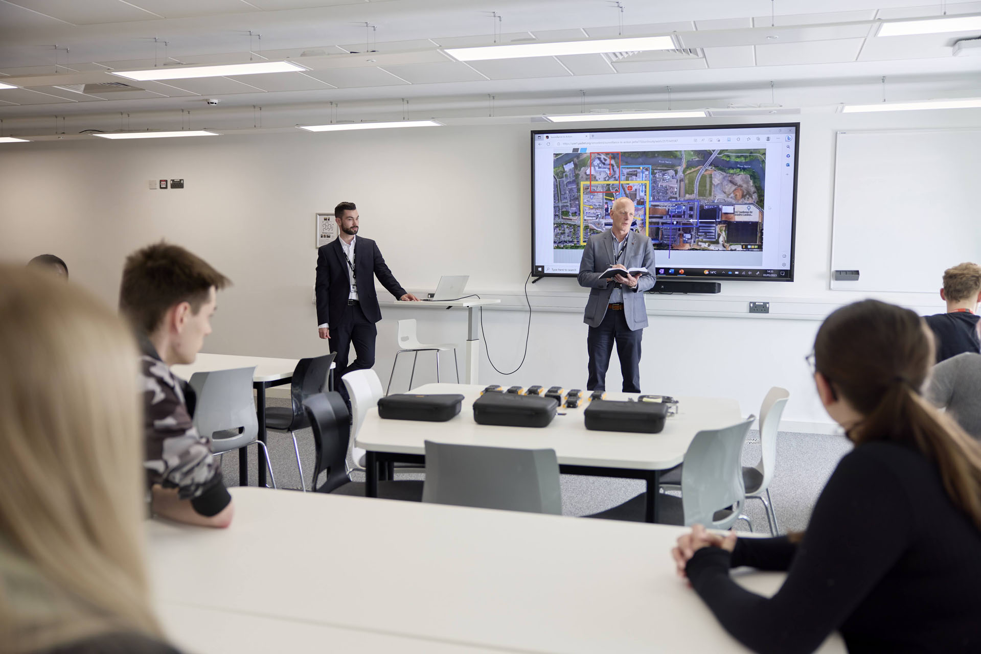 Two men dressed in smart clothing stand at the front of a classroom, next to a screen that is showing an annotated map of Waterside campus. They are facing groups of students sitting at tables.