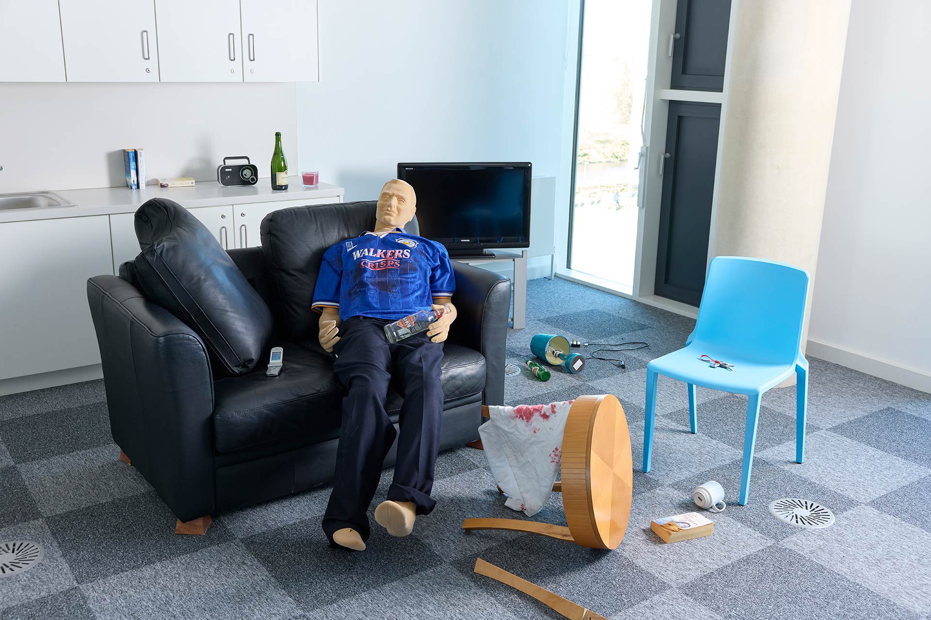 A clothed mannequin is lying on a sofa in a flat. The mannequin is wearing a blue football shirt and trousers, is lying next to a mobile phone, and is holding a bottle of vodka. The purpose-built flat has been made to look like a crime scene so there is fake blood on a towel, a broken table, a broken lamp, and various items strewn on the floor.