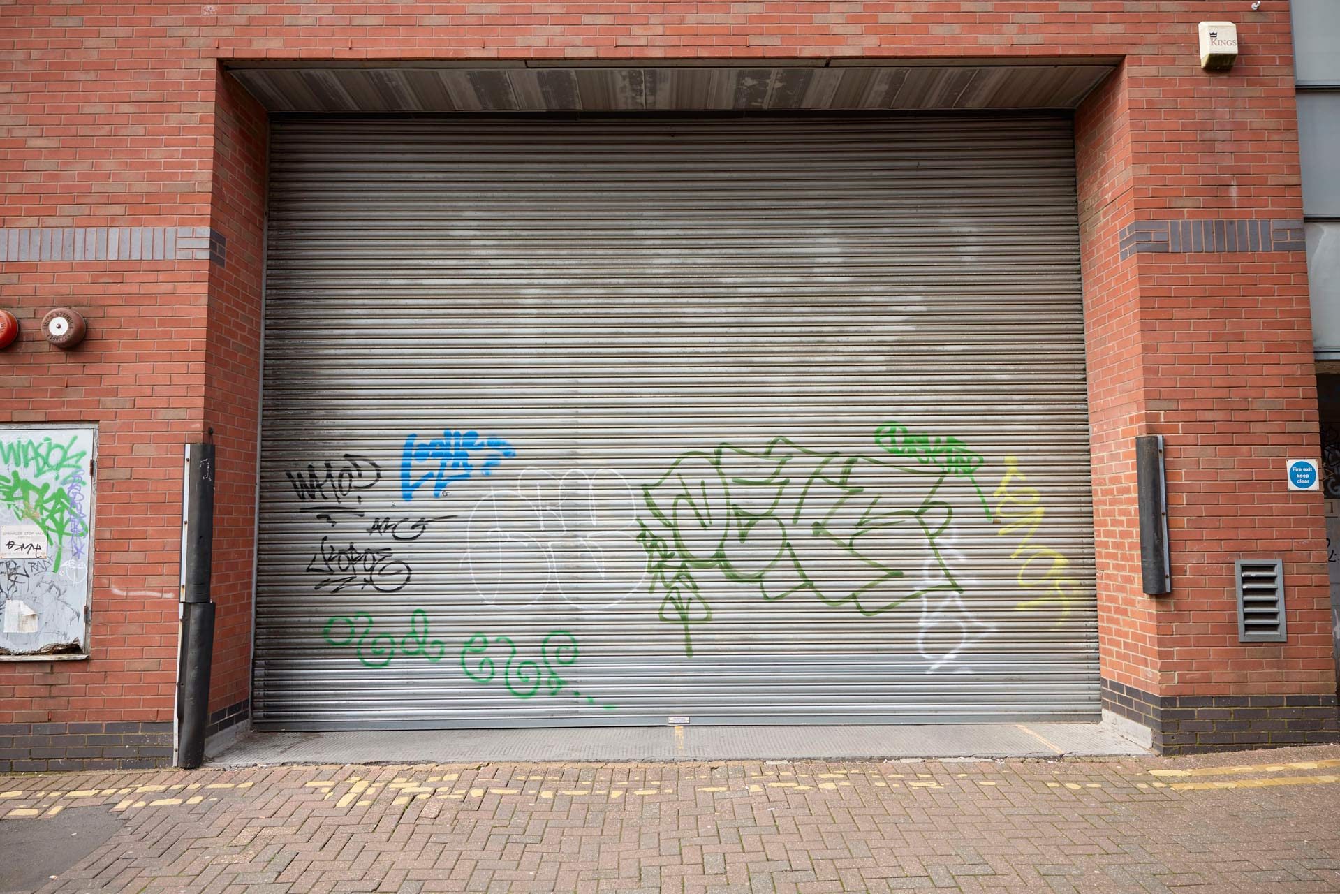A large set of closed metal shutters with various graffiti in blue, green, and black, surrounded by bricks.