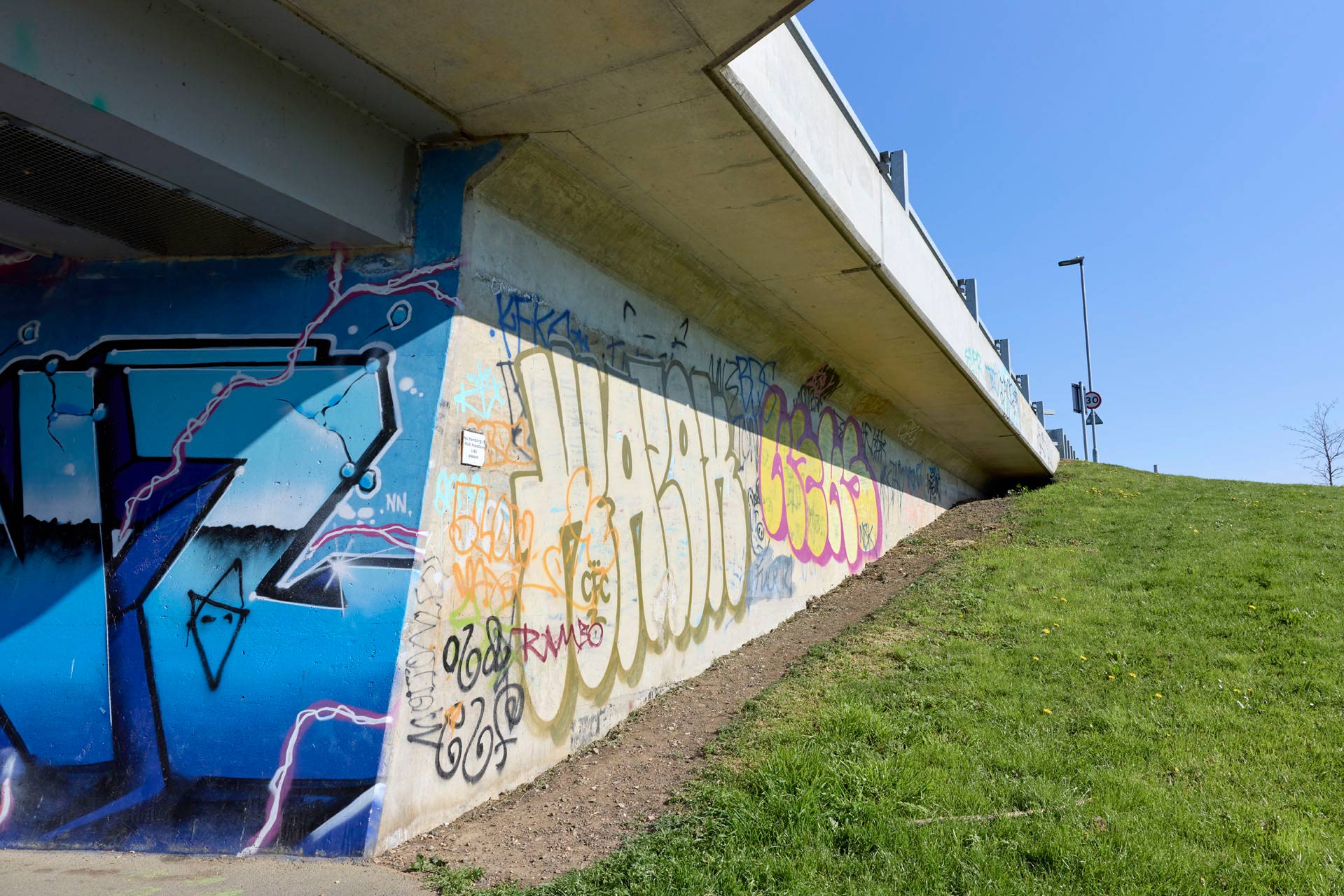 Large graffiti of various 'tagged' signatures and colours on an underpass wall under a road bridge, with a clear blue sky above.