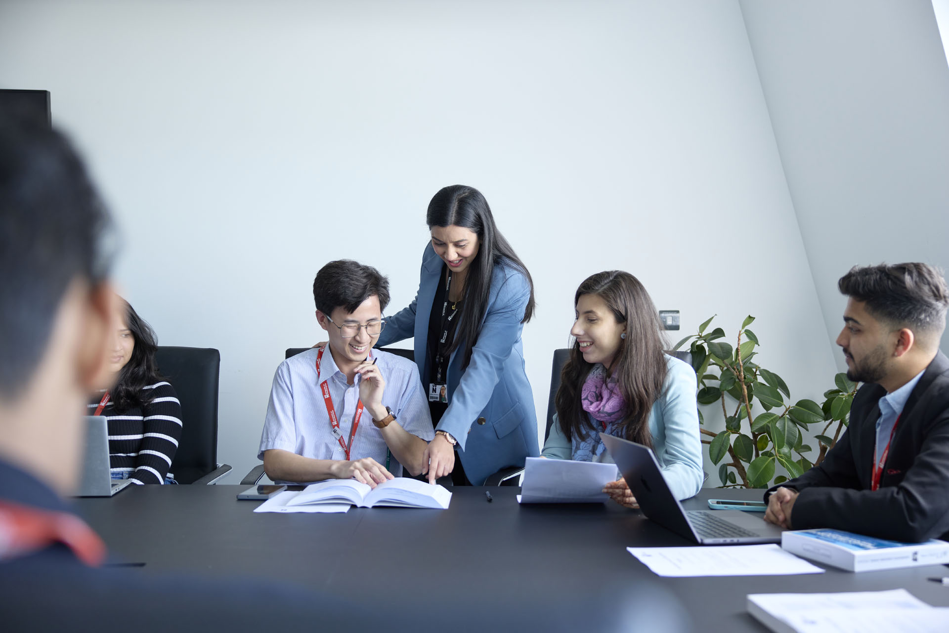 In an MBA class, a group of students are seated around a table, and behind them, an academic stands and points at a section in an open textbook during a discussion. On the table are open laptops, textbooks, and papers, and there is a large plant on the floor in the background.