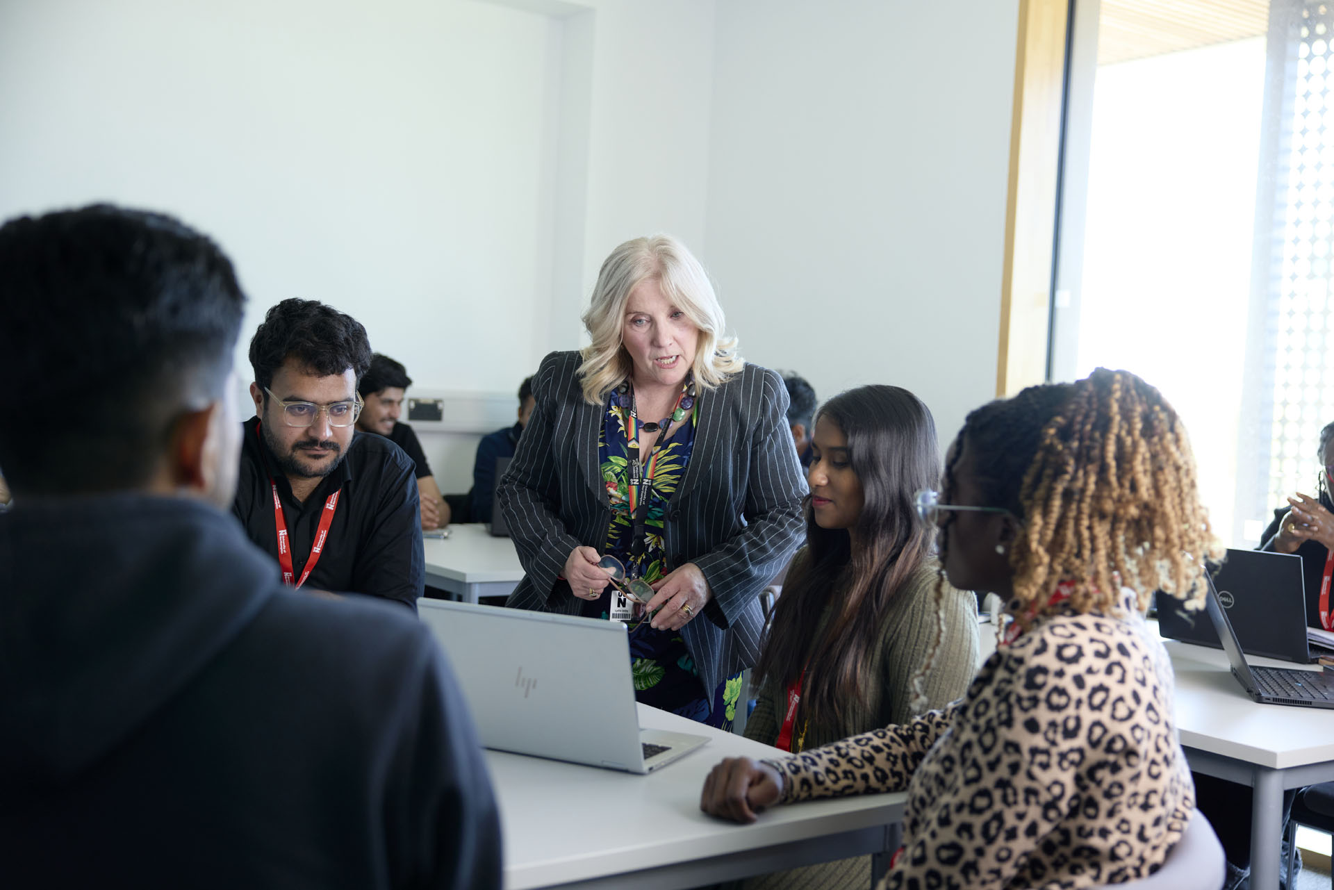 An academic discussing a topic with students during an MBA seminar. She is standing behind a table of seated students and they are all looking at an open laptop.