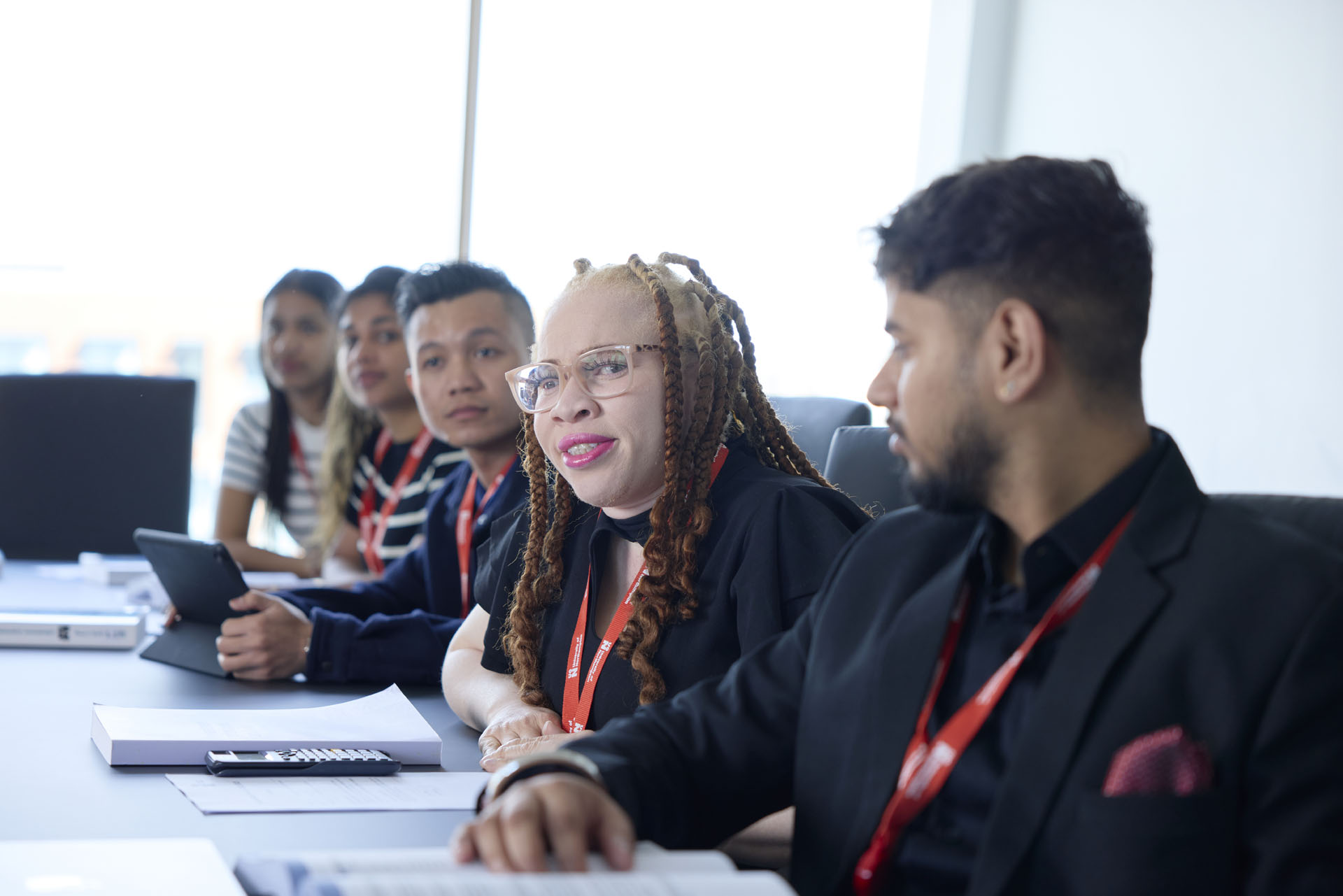 A group of students sit at a conference table during an MBA seminar. A woman is speaking while others listen. On the table are open laptops, textbooks, a calculator, and papers.