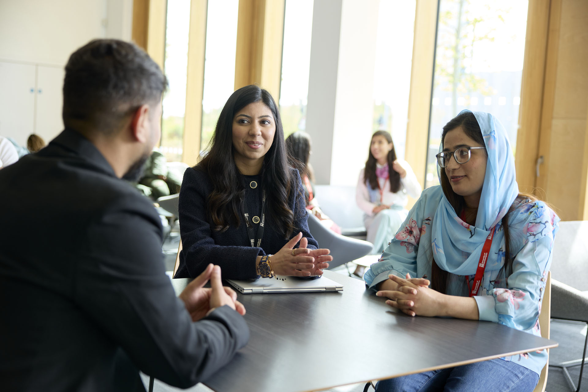 An academic discusses a topic with two MBA students, while seated at a table in a bright room. The academic is wearing a black jacket and is sitting between a woman wearing a blue hijab and a man in a dark suit.