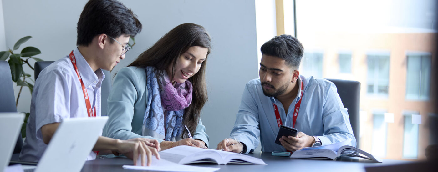 Three MBA students sitting at a table with open laptops and textbooks, working together during a class. A window in the background shows an outside view.