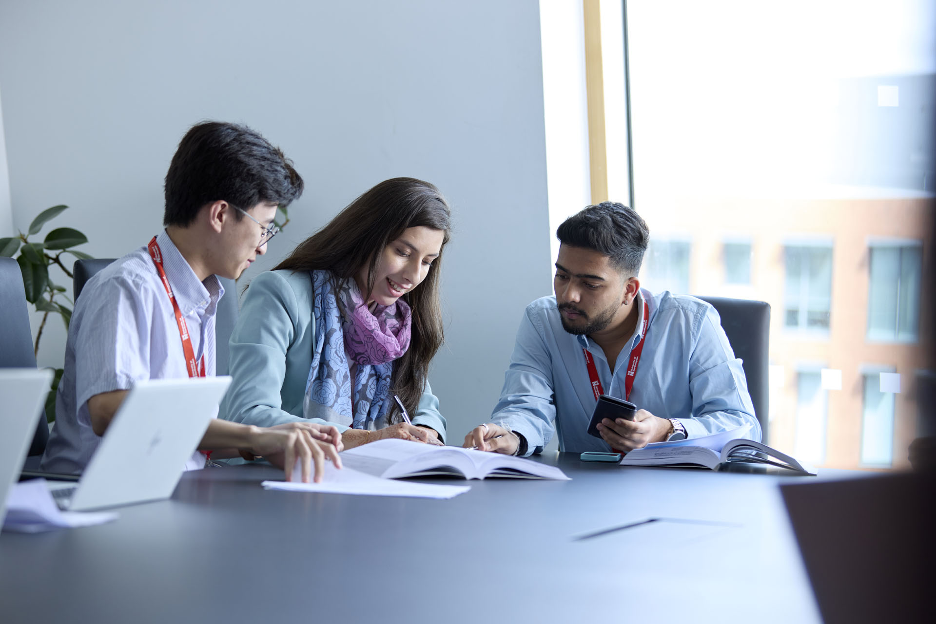 Three MBA students sitting at a table with open laptops and textbooks, working together during a class. A window in the background shows an outside view.