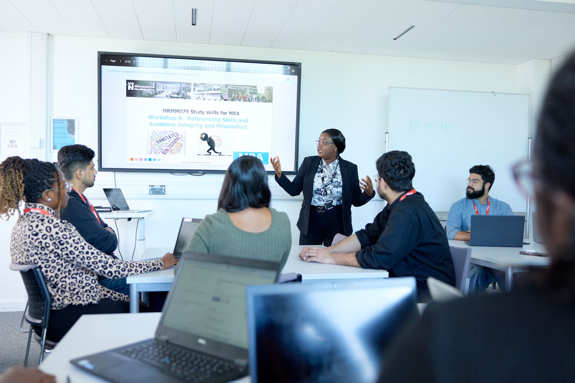 An academic presens to a class during an MBA study skills session. She is wearing a black suit and standing next to a display screen. Around her, students are seated at tables with open laptops.