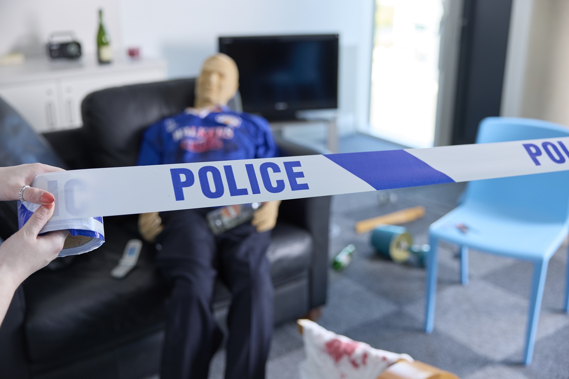 In the foreground, a pair of hands pulls blue and white POLICE tape across a fake crime scene. In the background, a mannequin on a couch surrounded by scattered items is blurred.