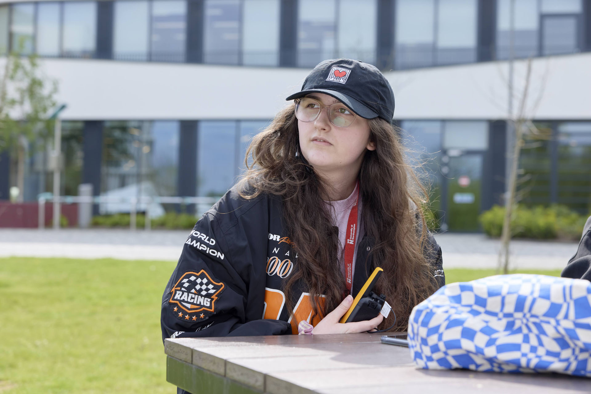Student with long hair, wearing a baseball cap and glasses sits at a picnic table. She is holding a yellow walkie-talkie close to her body. Visible on the table is a bag and a mobile phone.