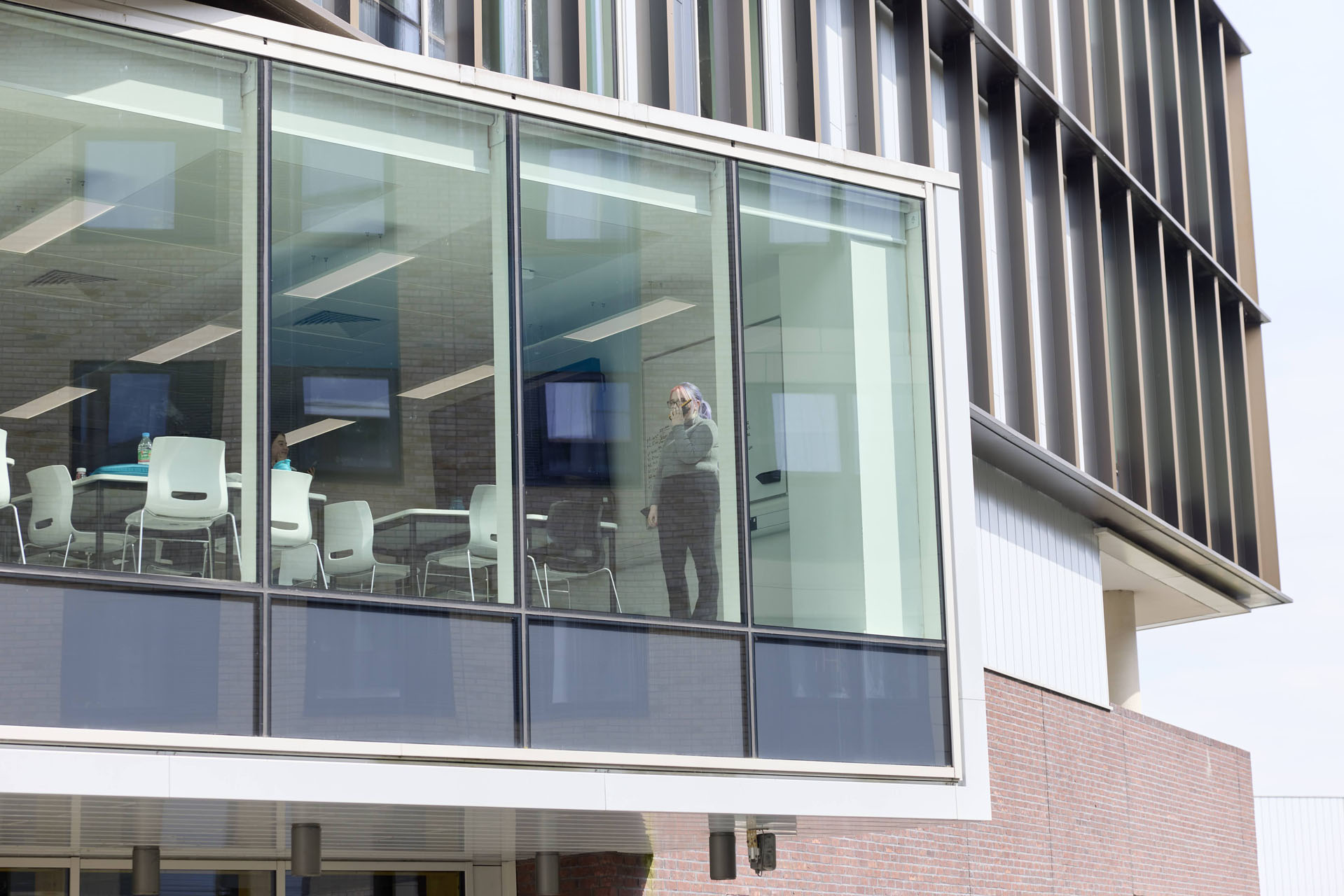 A student stands inside a modern building with large glass windows, alongside empty chairs and tables. She is speaking into a mobile radio while looking out of the floor-to-ceiling window.