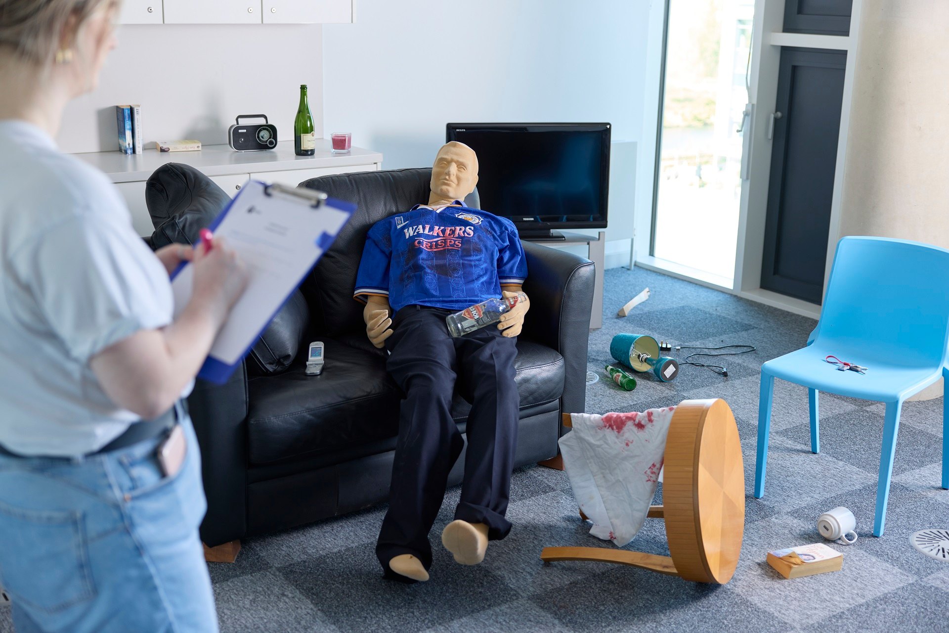 Professional Policing student observing a mock crime scene in UON's purpose-built flat while taking notes on a clipboard. In front of them is a clothed mannequin lying on a sofa. Next to the sofa is a towel with fake blood on a broken table, a broken lamp, and various items strewn across the floor.