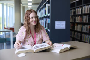 A student sits at a table in the library, with shelves full of books behind them. She has curly hair and is looking through one of the two open textbooks that are on the table.