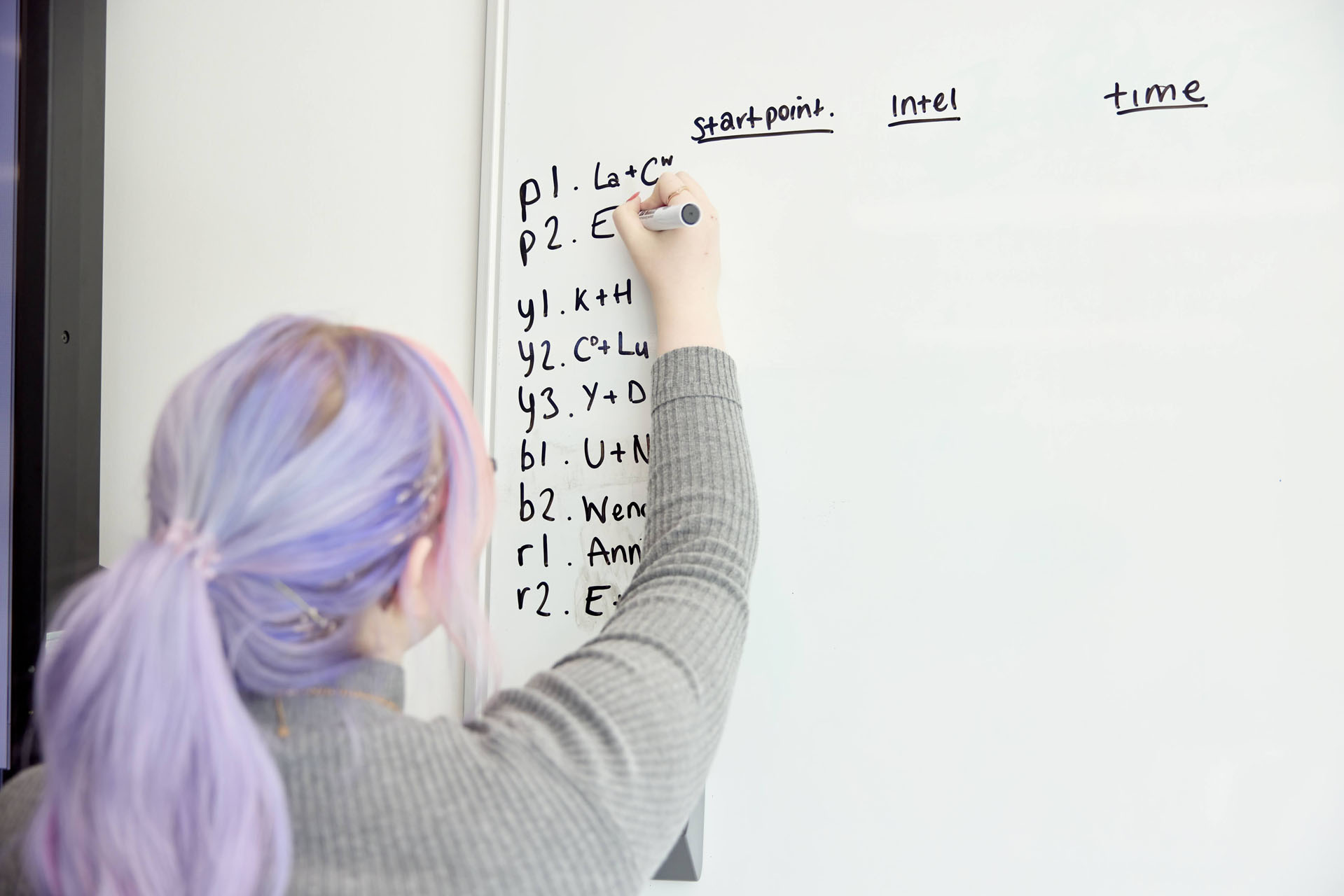 A student with pastel purple and pink hair and a light grey jumper uses a marker pen to write location and time data on a whiteboard.