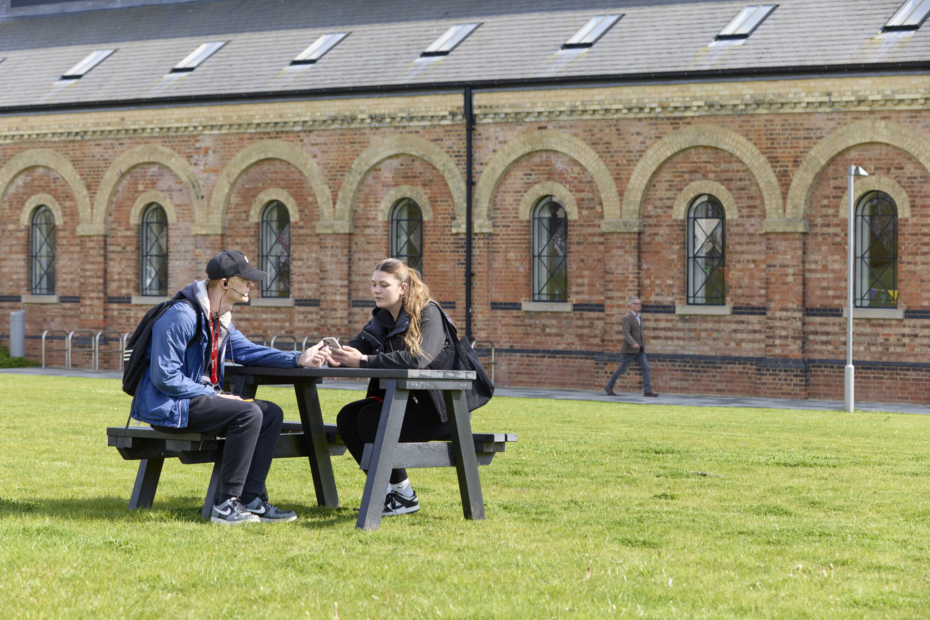 Two students sit at a picnic table on a grassy area outside a brick building. They are wearing radios and microphones, and observing a man wearing glasses and jeans who is walking down a path behind them.