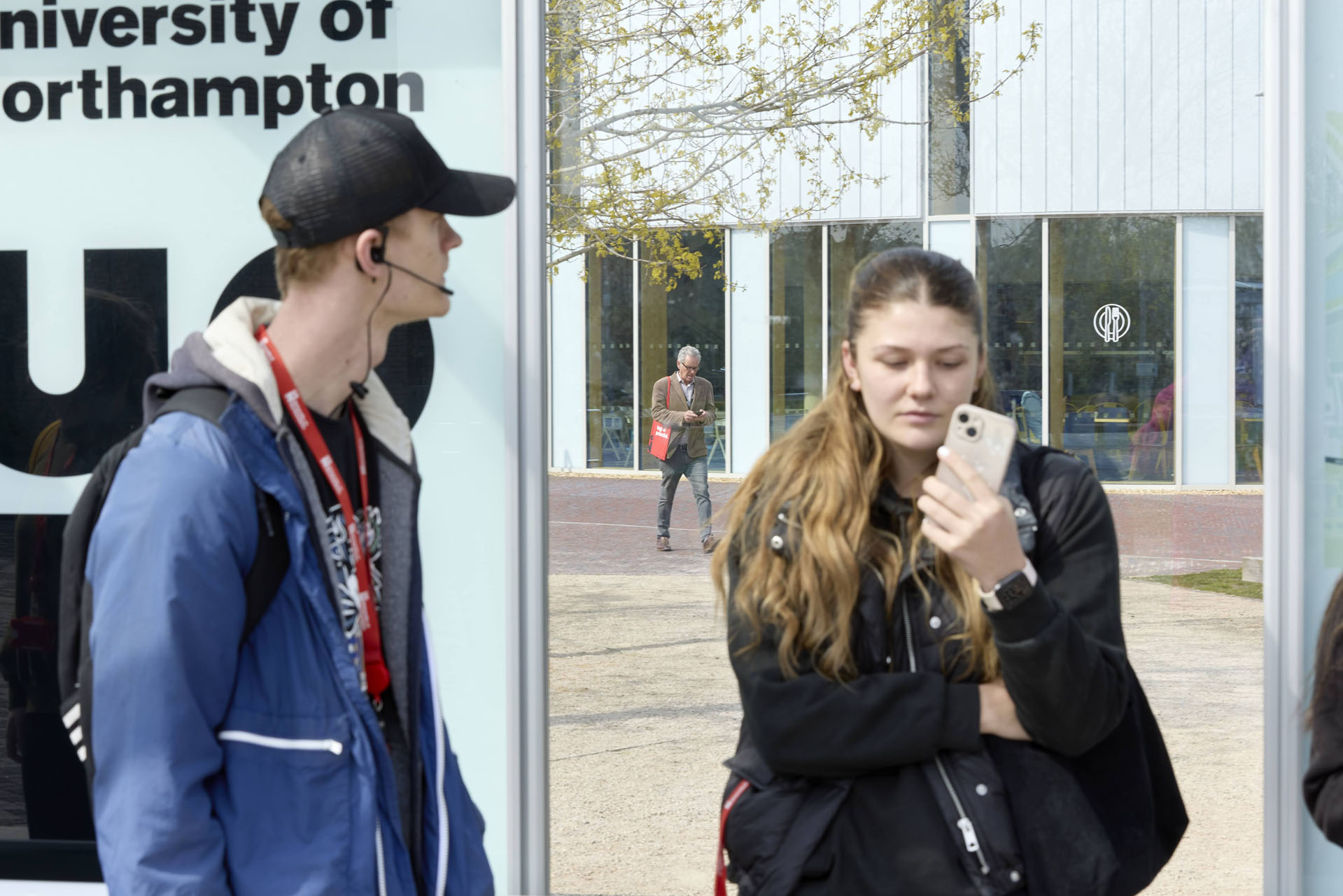 Two students stand at a bus stop; one is looking down at a mobile phone, while the other who is wearing a baseball cap and a headset is looking to the side. In the background, a man is walking past a glass-fronted building while carrying a red cloth bag on his shoulder.