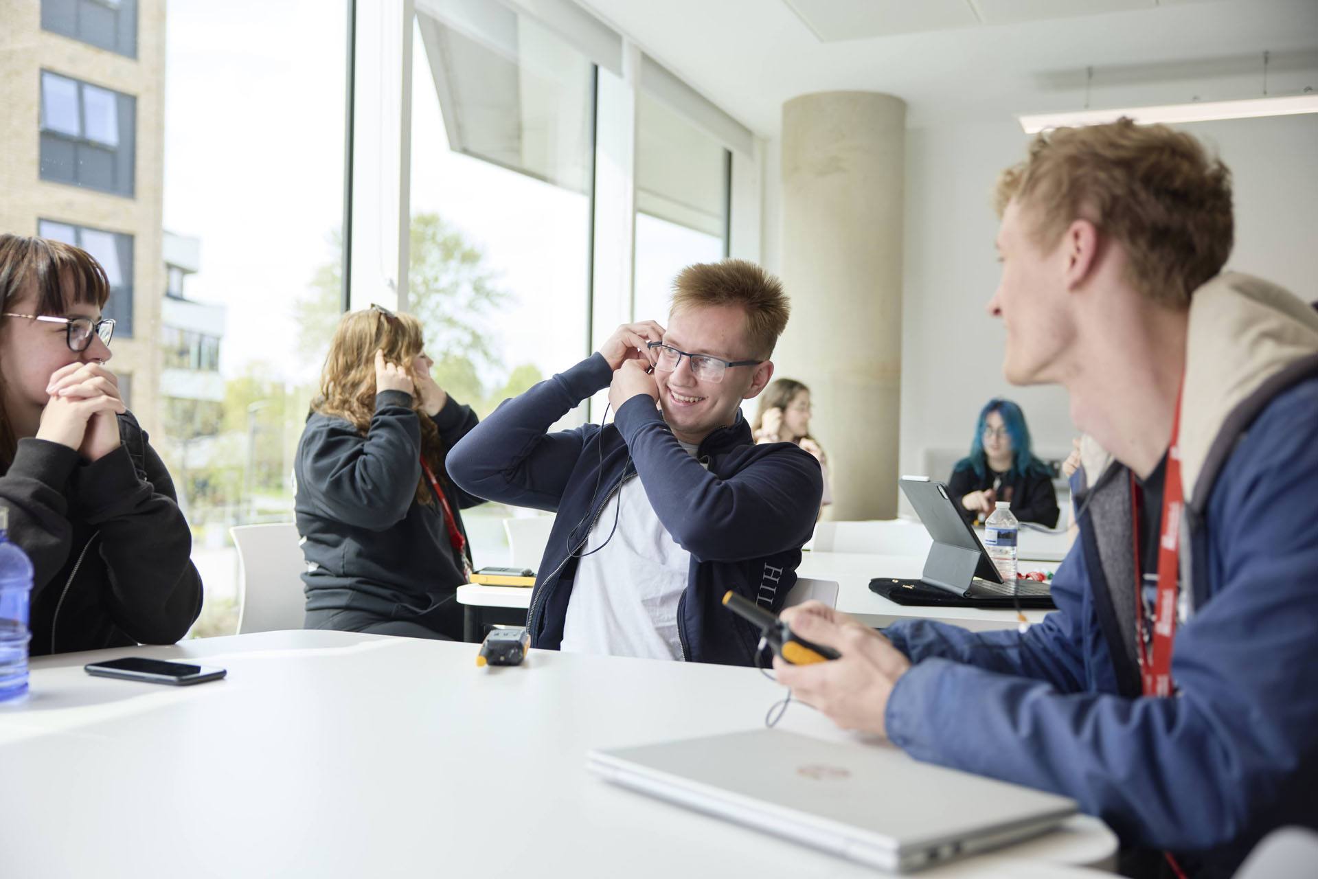 A group of students in a bright classroom setting are seated at tables. Some are holding radios to their ears to test them. On the tables are laptops, mobile phones and water bottles.
