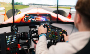 Person simulates driving a race car using a steering wheel in front of a multi-screen setup displaying a racing game.